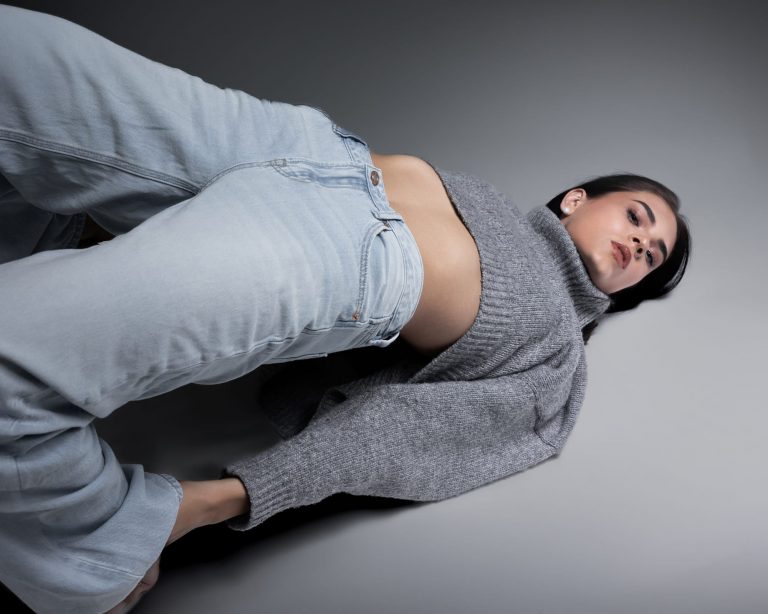 Model mit Jeans liegt auf grauem Studiohintergrund und blickt nach oben in die Kamera, Sedcardshooting Vorarlberg