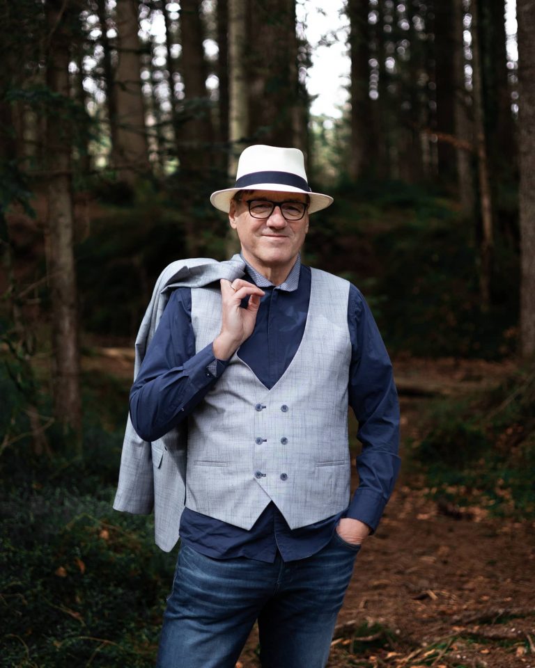 Mann mit Hut und Weste posiert entspannt im Wald, Outdoor Portrait Vorarlberg.