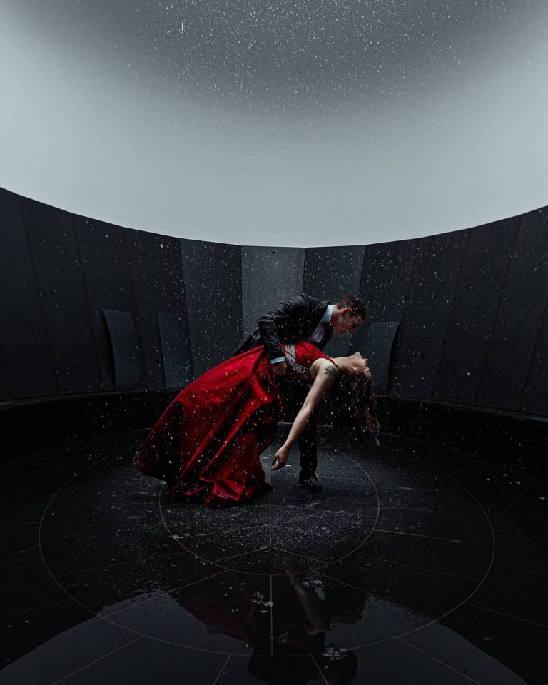 Elegantes Tanzpaar in rotem Kleid und Anzug posiert im Regen bei künstlerischem Fotoshooting in Vorarlberg.