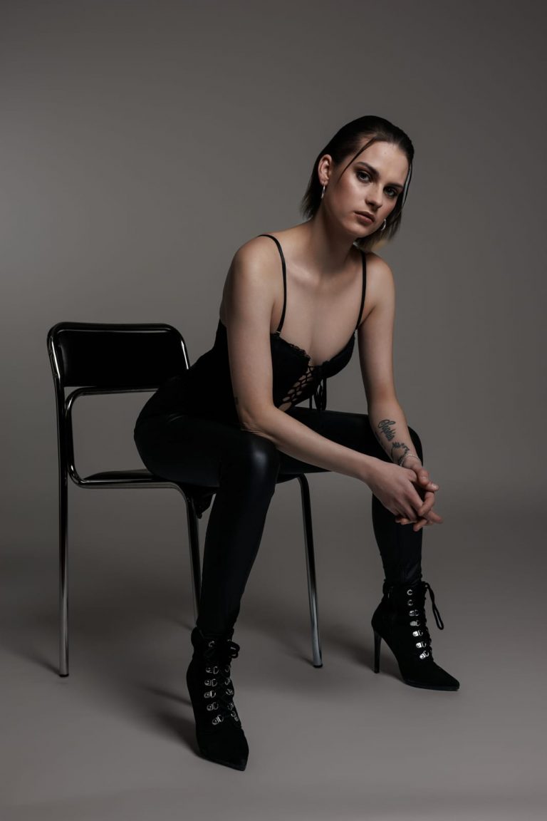 Selbstbewusstes weibliches Model in schwarzem Spitzen-Body und Lederhose auf einem Stuhl in dramatischem Studiolicht. Model Shooting, Model Fotoshooting Vorarlberg, Sedcardshooting Vorarlberg, Sedcardfotos.