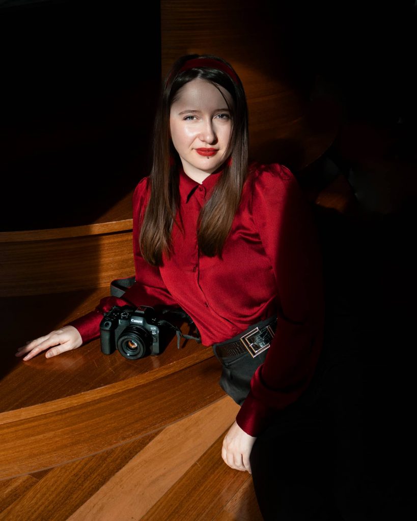 Portrait von Viktoria Jäger, Fotografin in Vorarlberg, sitzend auf einer hölzernen Wendeltreppe mit roter Bluse und Kamera in der Hand, eingefangen im natürlichen Licht – spezialisiert auf professionelle Fotoshootings in Vorarlberg