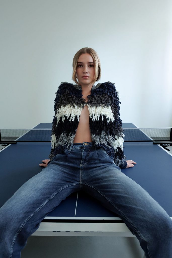 Geometrische Fotoaufnahme, Model sitzt auf einem blauen Tischtennistisch mit blau, grauer Jacke, Fashion Fotografie Vorarlberg