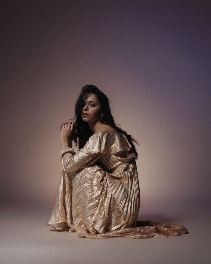 Model mit goldenem Kleid sitzt im Fotostudio und schaut in die Kamera, der Hintergrund ist violet, rosa beleuchtet, Mode Fotograf Vorarlberg
