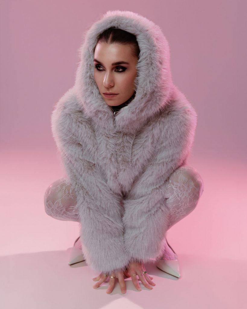 Model im Pelzmantel sitzt im Studio mit rosa Hintergrund, Editorial Fashion Foto Vorarlberg